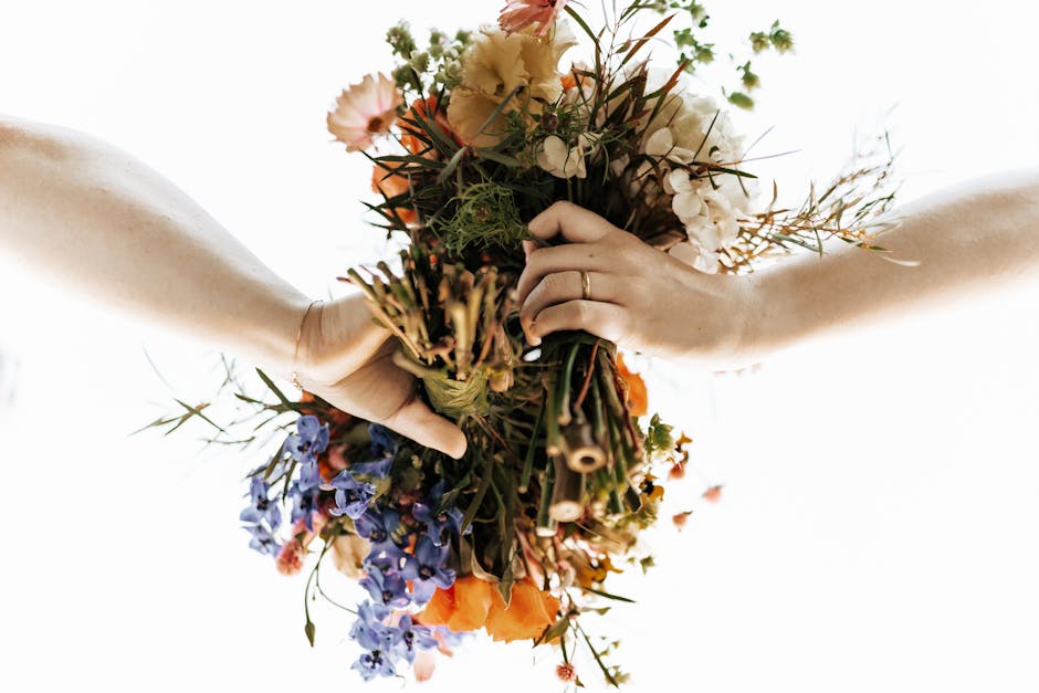 A pair of hands holding a vibrant floral bouquet featuring a mix of flowers in shades of orange, white, purple, and blue, with green foliage and stems. The arrangement appears fresh, with petals that 