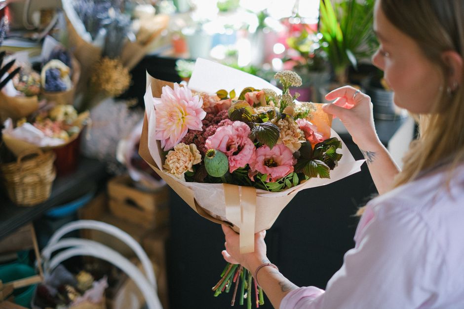 A woman holding a large bouquet of fresh flowers indoors, with a mix of pink and peach blooms including large dahlias, lisianthus, and small filler flowers, wrapped in cream and brown paper with a sil