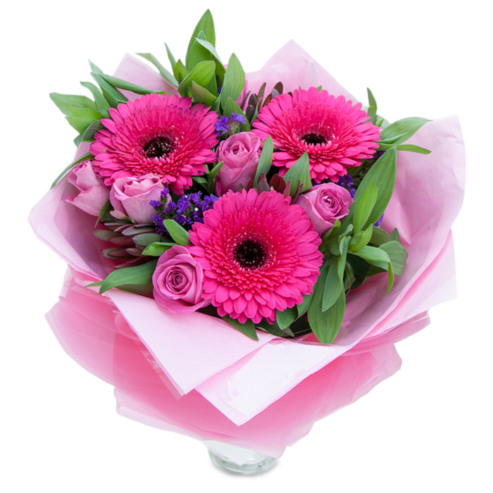 I see this arrangement as a tender, playful portrait in pink: three vivid fuchsia Gerbera daisies open like small suns, their many petals catching light and creating a bold, joyful heart to the bouquet. Around them, lavender-pink roses curl inwards with the promise of unfolding, their dusty-rose tones softening the intensity of the Gerberas and lending the composition a romantic hush. Tiny clusters of purple statice or limonium provide a lively, textured punctuation between the larger flowers, their papery florets recalling summer meadows along the edge of Virginia Water Lake. The foliage-lush, green, and varied-resembles salal and eucalyptus, giving depth and a cool, slightly aromatic lift when the stems are handled. The flowers are cradled in an inner layer of soft pale pink tissue and wrapped over with a sheer fuchsia-pink cellophane that shimmers faintly where the light hits its gentle crinkles; the wrapping is folded with care, a quiet sign of professional craft. The rim of a clear glass vase is visible at the base, suggesting immediacy and readiness to display on a dining table after a stroll around the lake or a visit to the nearby village shops. In my mind the scent is fresh and understated-roses offering a gentle perfume, Gerberas adding a green sweetness-making this arrangement fitting for a birthday, a thank-you, or an intimate celebration by the ponds at Virginia Water. Described for someone who relies on words, the bouquet balances confident, bright focal blooms with tender accents, textured fillers, and verdant greenery in a finished gift-ready presentation.